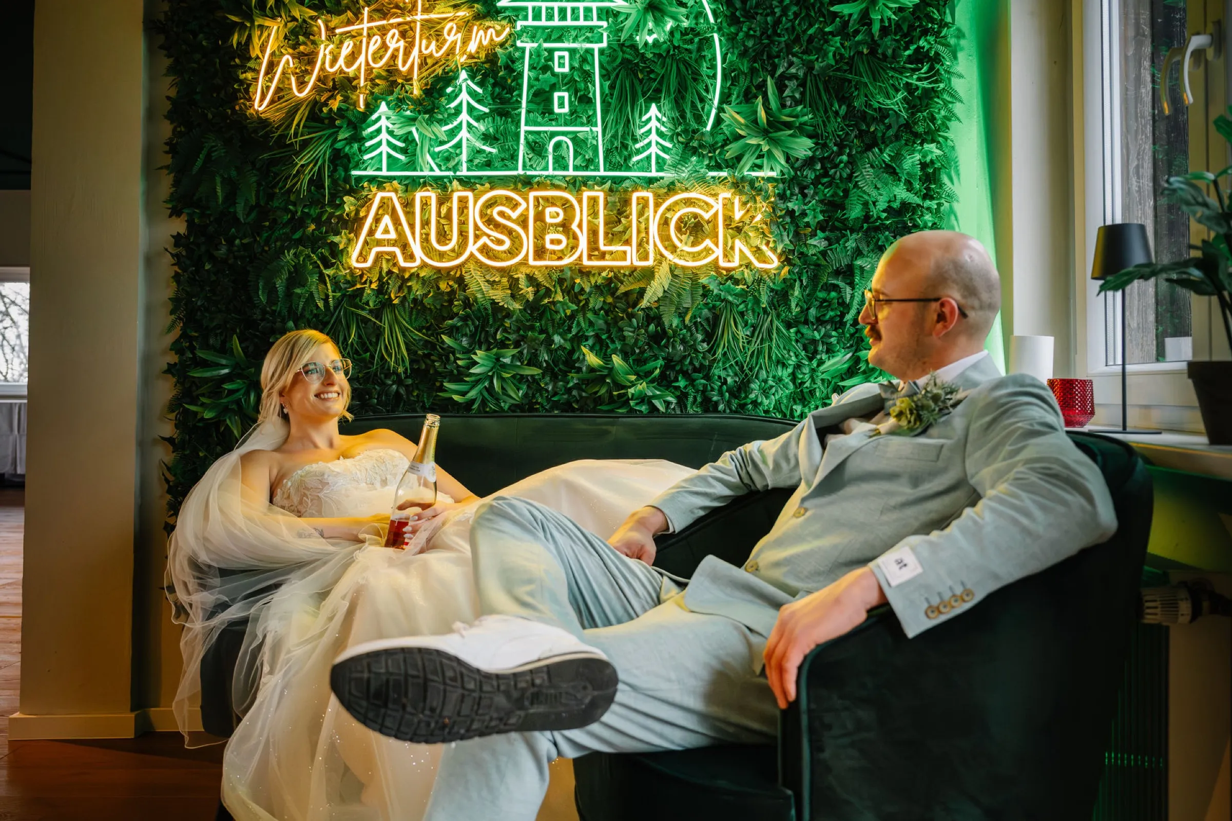 Brautpaar entspannt auf dem Sofa vor der Neon-Mooswand